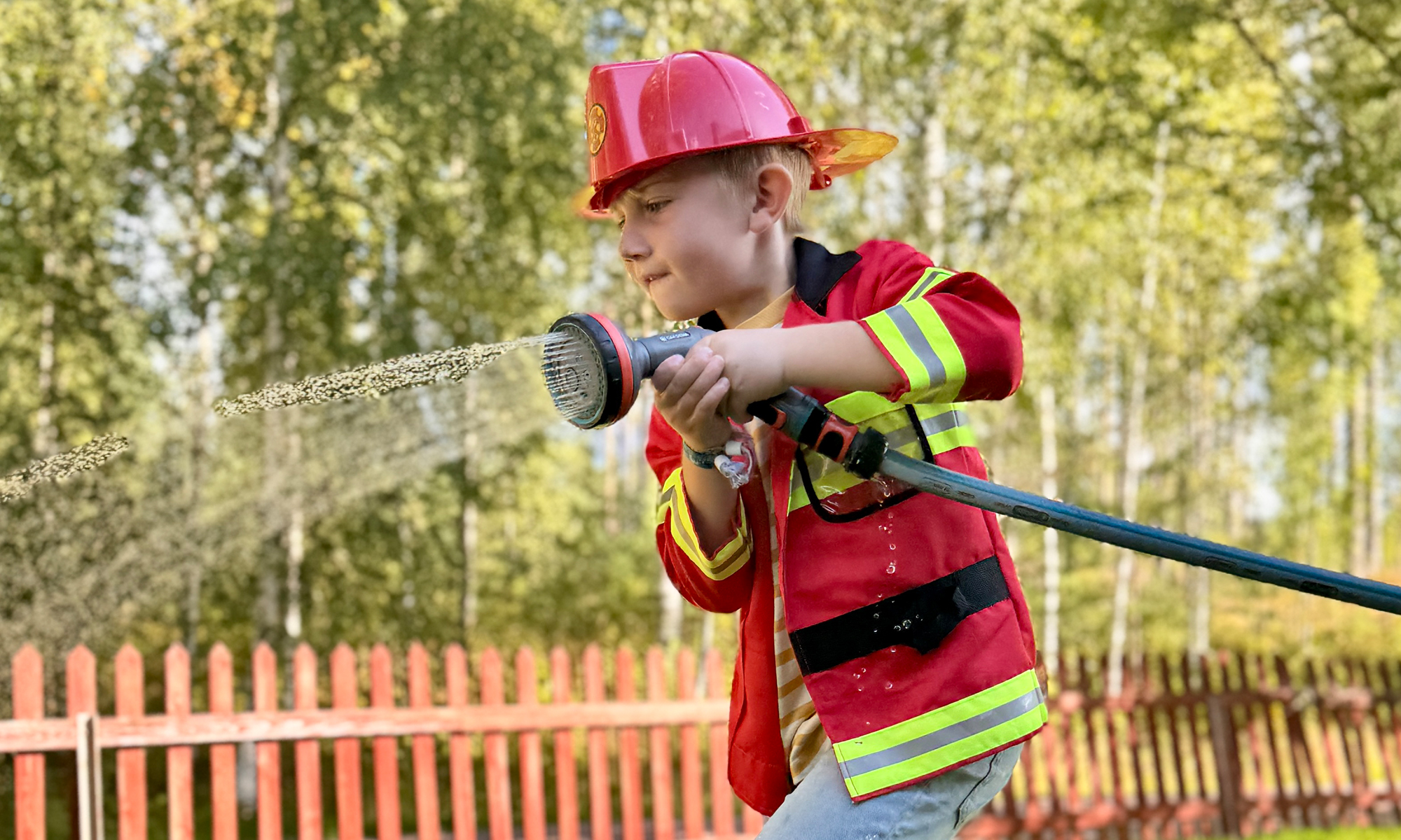 En barn utklädd till brandman leker utomhus med en vattenslang.