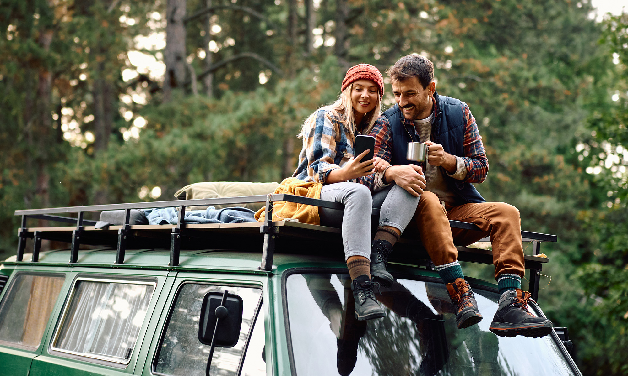 En kvinna och en man sitter och fikar på taket på sin husbil i en skog