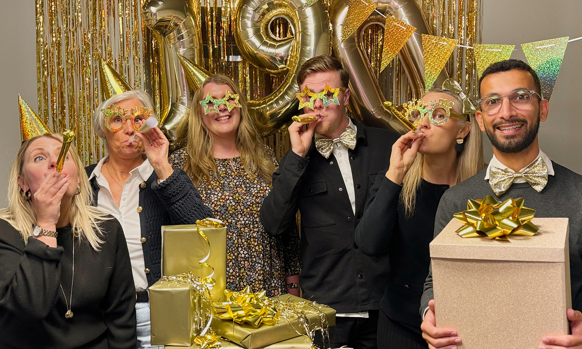 Personal som firar och har festliga guldattiraljer på sig. I bilden finns även flera paket, vimplar, ballonger och draperi i guld.